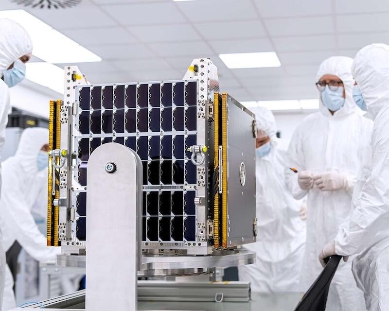 engineers in white cleanroom suits inspect a compact satellite