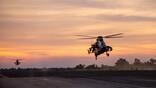two military helicopters fly low over a runway at sunset