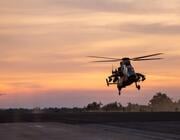 two military helicopters fly low over a runway at sunset