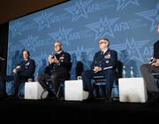 four U.S. military officers in chairs next to suited man by podium at conference